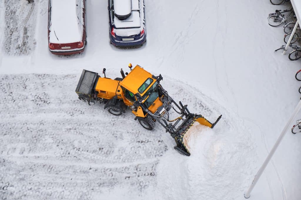 Keltainen auraaja puhdistaa lunta parkkialueella ylhäältä kuvattuna.